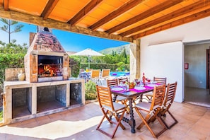 An outdoor patio with a brick oven, wooden table, and chairs, overlooking a pool and greenery.