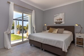 A bedroom with a large bed, a sliding glass door to a balcony, a nightstand with a lamp, and a painting on the wall.