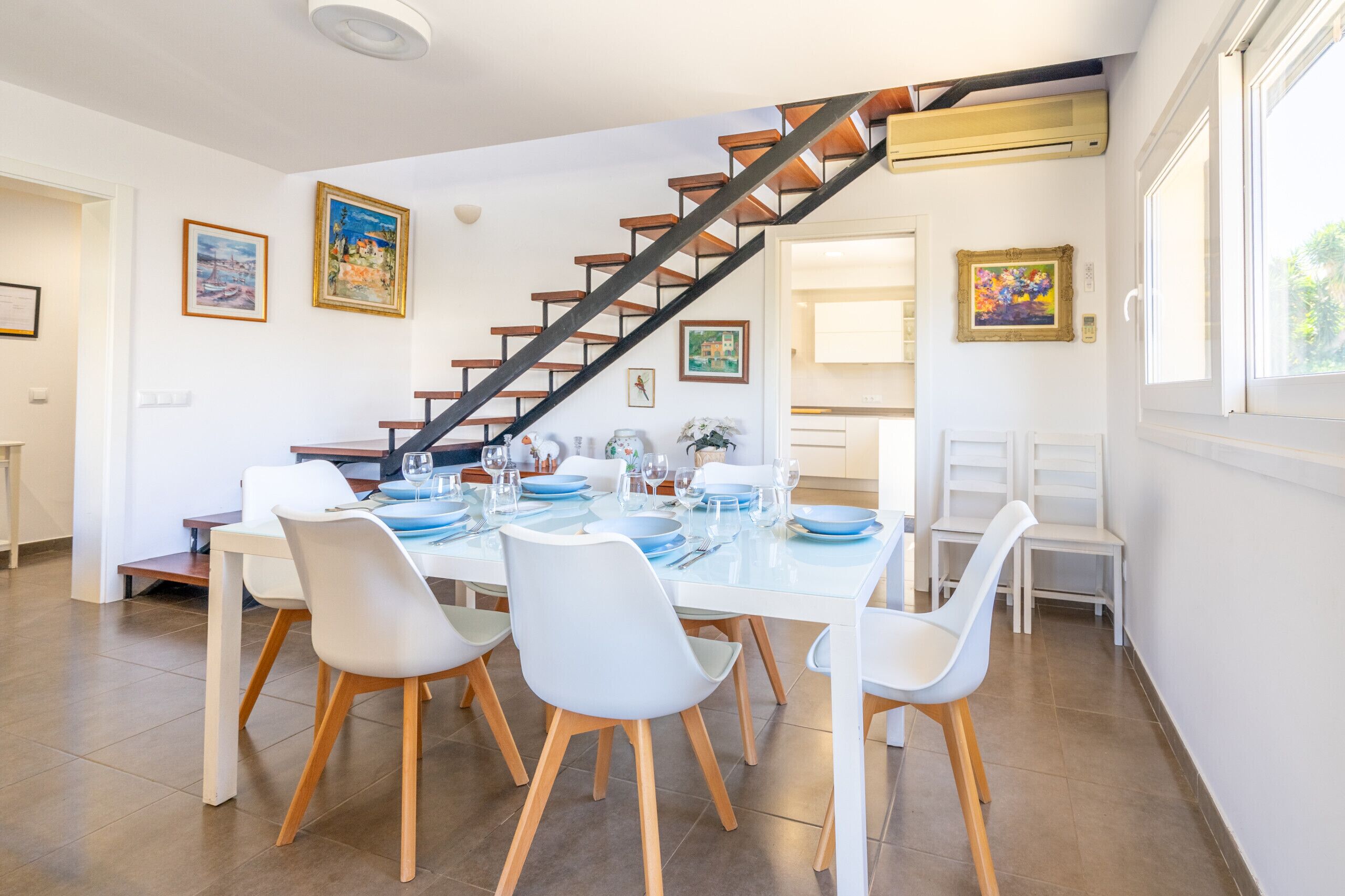 A dining area with a white table set for four, a staircase with wooden steps, and framed artwork on the walls.