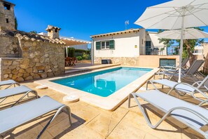 A swimming pool with lounge chairs and an umbrella, stone walls, and a small building with a window.