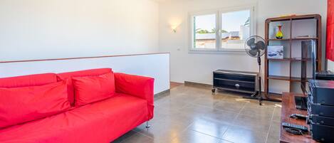 A room with a red sofa, a fan, a wooden shelf with a vase, and a window with a view of the outside.