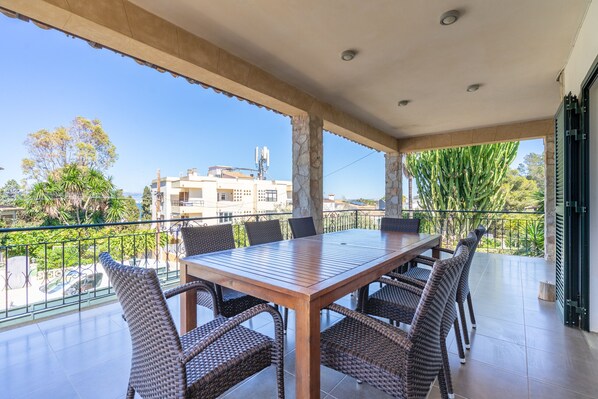 A covered outdoor dining area with a table and chairs, a view of buildings and greenery, and a balcony railing.