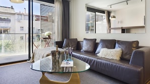 Un salon moderne avec une table basse en verre, un canapé en cuir et de grandes fenêtres donnant sur des immeubles et des arbres.
