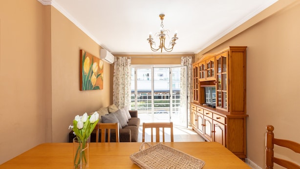 A dining room with a wooden table, chairs, a vase of flowers, a chandelier, and a view of the outdoors through a glass door. - CARLOS V Apartment (Salou)