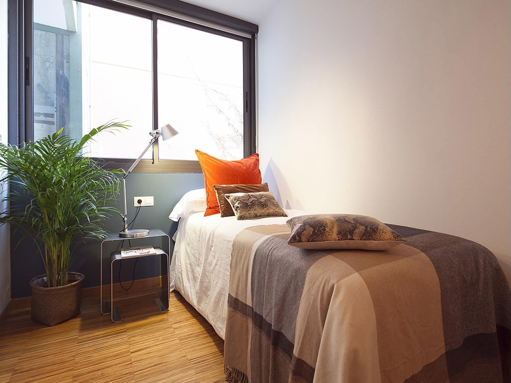 Un dormitorio con cama, mesita de noche, lámpara, planta en maceta y ventana.