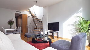 Un salon moderne avec un canapé blanc, un fauteuil gris, une table basse en verre et un téléviseur posé sur un meuble. Un escalier avec des marches en bois et des rampes en métal mène à une autre pièce.