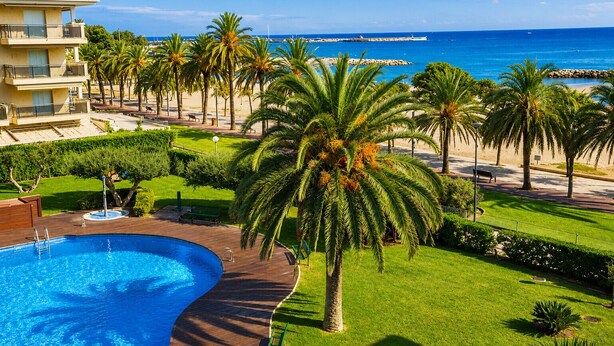 A poolside view with a clear blue pool, palm trees, and a distant view of the sea and a pier. - GOLF SANT JORDI 2 Apartment (Cambrils)