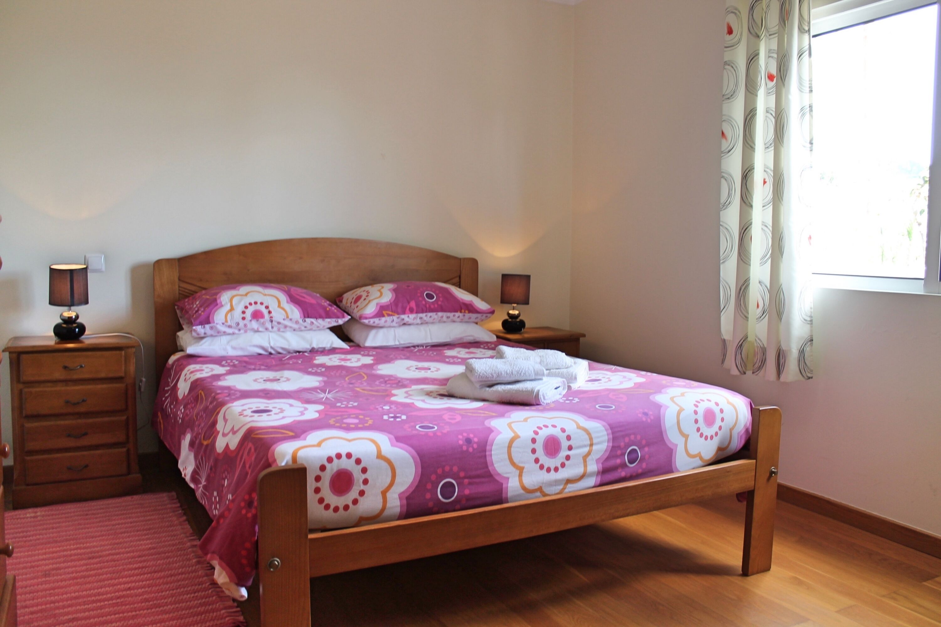 Une chambre à coucher avec un lit en bois, deux tables de chevet avec des lampes, une fenêtre avec des rideaux et un tapis rouge.