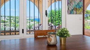 Un comedor con vista al mar a través de puertas metálicas decorativas, una mesa de madera, una planta en maceta y un cuadro abstracto y colorido en la pared.