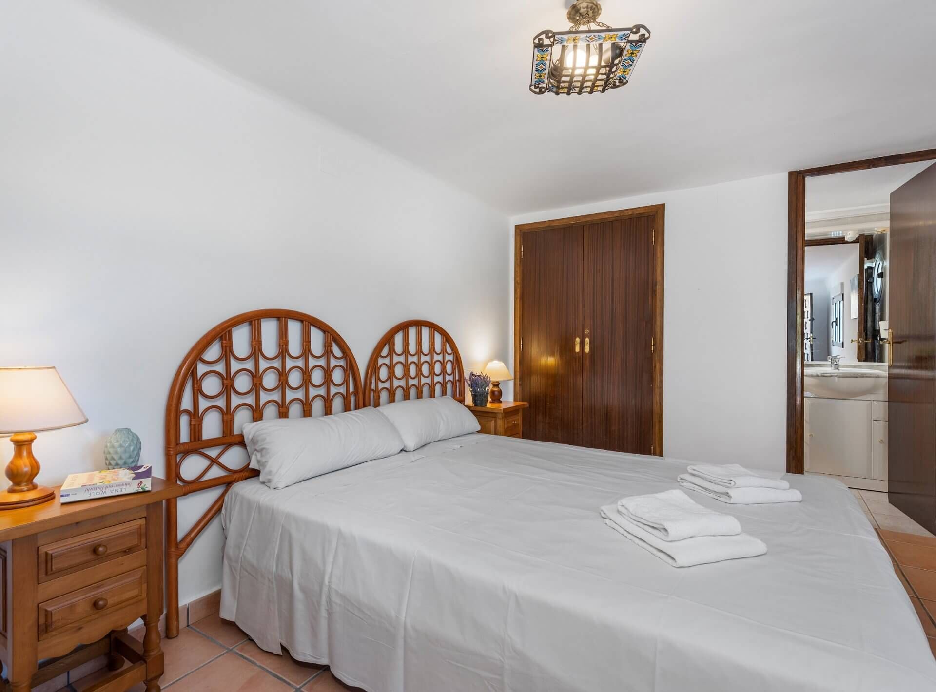 A neatly made bed with white linens, a wooden headboard, a bedside lamp, and a nightstand with a book and a decorative item.