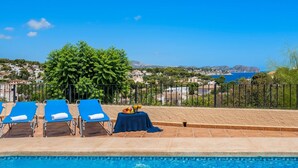 Un área junto a la piscina con sillones reclinables, una mesa con fruta y vistas al mar y a las colinas.