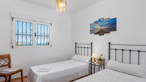 Una habitación con dos camas, una ventana con barrotes, un cuadro de un atardecer y una lámpara colgante.