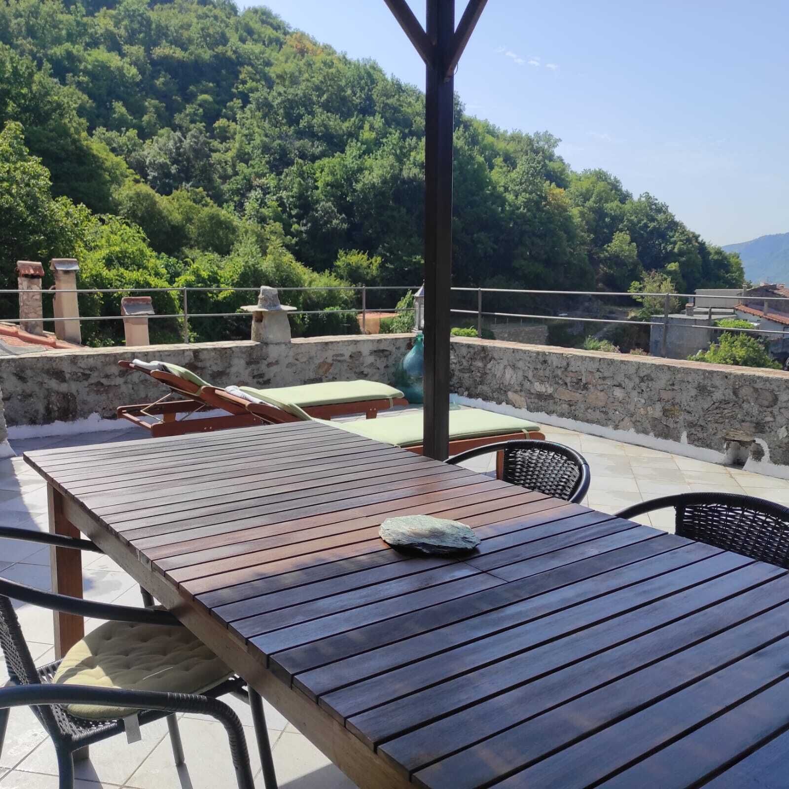 Una mesa de madera con una piedra encima, rodeada de sillas de mimbre, ubicada en una terraza con muro de piedra y vista a una colina boscosa.