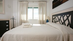 A neatly made bed with a white duvet and two folded towels, a bedside lamp, and a view of the outdoors through sheer curtains.