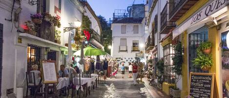 A narrow street with shops and outdoor seating, stone pavement, and balconies with plants.