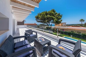 A balcony with wicker furniture, a glass railing, and a view of a swimming pool and trees.