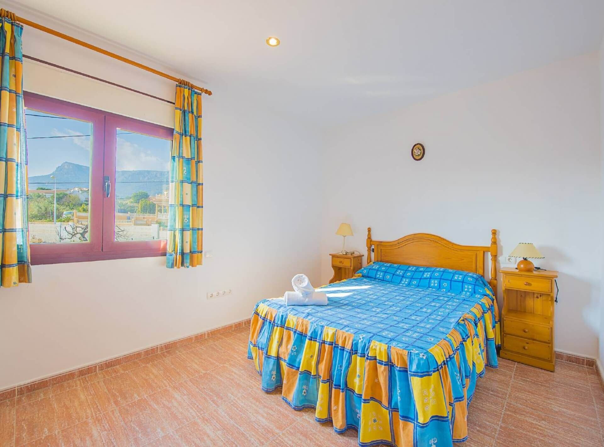A bedroom with a bed, two bedside tables, a window with curtains, and a view of the outside.