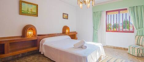 A hotel room with a large bed, a chandelier, a window with curtains, and a painting on the wall.
