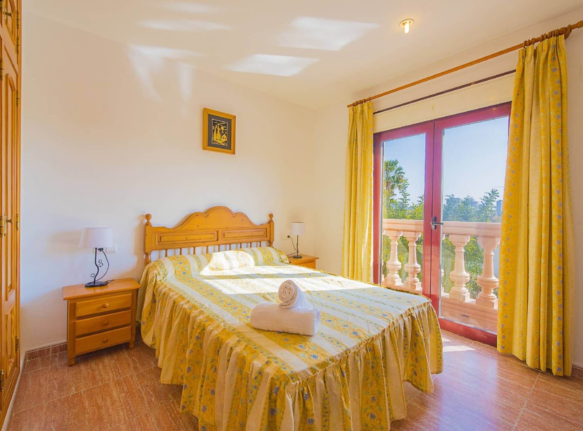 A hotel room with a bed, two bedside tables, a window with yellow curtains, and a balcony with a railing.
