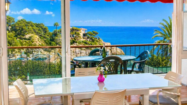Un balcon avec une table et des chaises, donnant sur un paysage côtier avec des arbres et la mer.