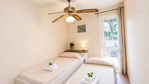 Une chambre avec deux lits, un ventilateur de plafond, une fenêtre avec des rideaux et une petite table sur laquelle est posée une plante.