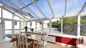 A conservatory with a wooden table and white chairs, large windows, and a view of the outside.