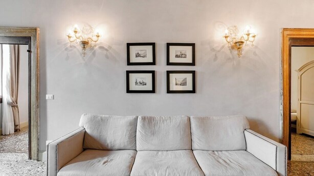 A neutral-toned sofa with a cushioned backrest and armrests, positioned against a wall with four framed pictures and two wall-mounted light fixtures. - Palazzina Canal Apartment (Venezia)