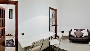 Une pièce avec un bureau blanc, une chaise, un canapé avec des coussins, une porte en bois et un miroir.