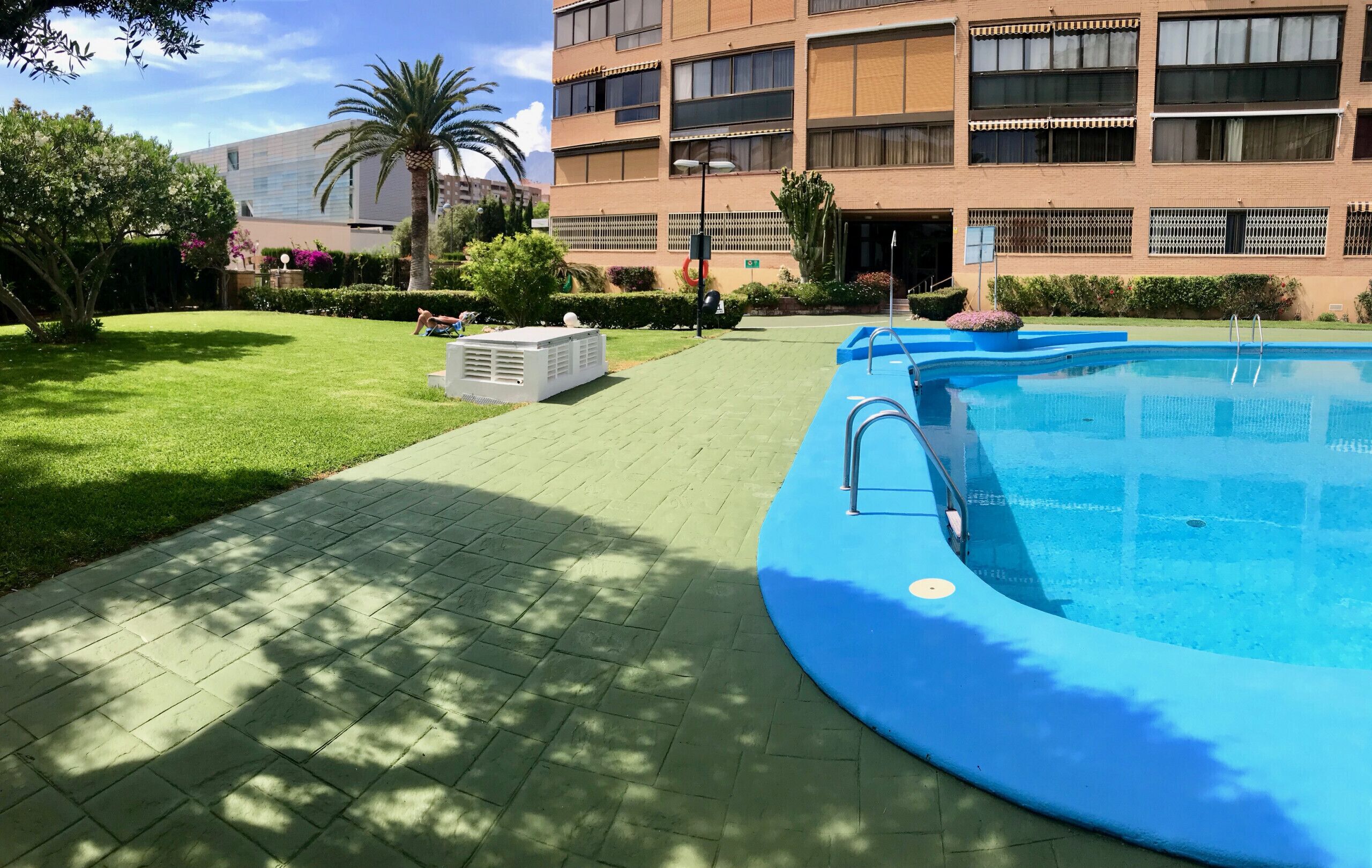 A pool area with a blue pool, a brick pathway, and a building with multiple windows in the background.