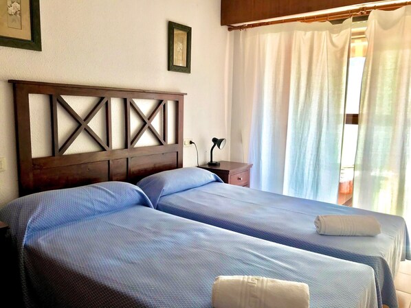 A room with two beds, a wooden headboard, a nightstand, a lamp, and a window with white curtains. - ALBAIDA BLUELINE R015 (Benidorm)