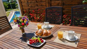 Une table en bois avec des aliments pour le petit-déjeuner, un vase de fleurs et une vue sur une piscine et la verdure environnante.