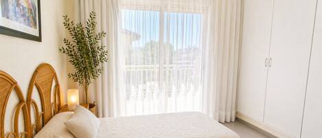 A bedroom with two beds, a nightstand with a lamp, a potted plant, and a view of a balcony.