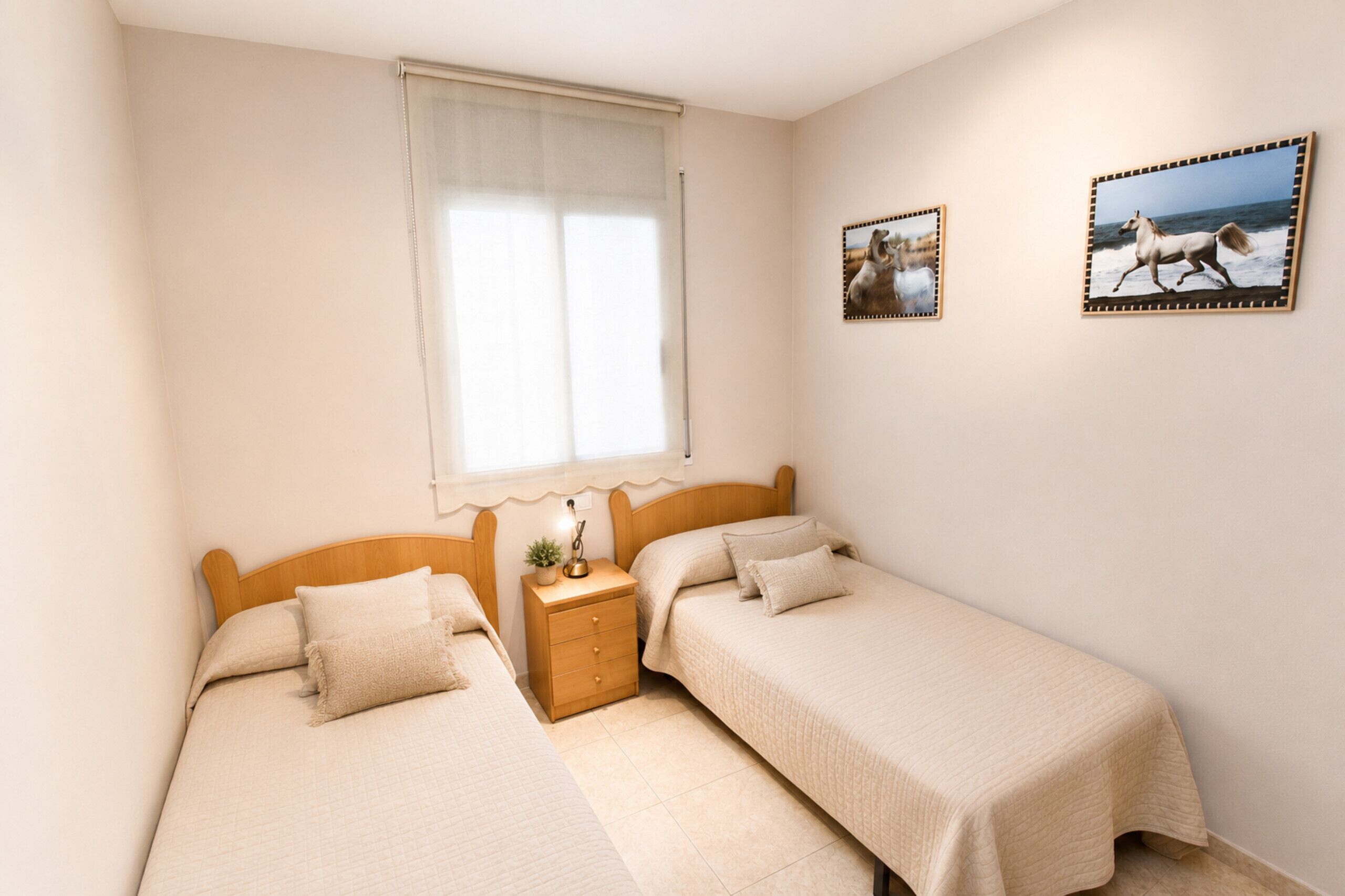 A bedroom with two beds, a nightstand, a window with curtains, and two framed pictures on the wall.