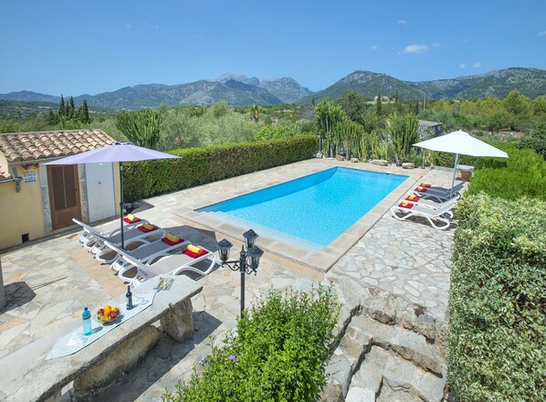 A swimming pool with lounge chairs and umbrellas, surrounded by stone tiles and greenery, with mountains in the background.