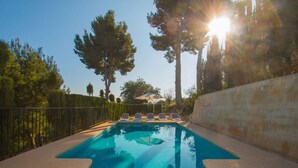 A swimming pool with clear blue water, surrounded by a low stone wall and a metal fence. There are lounge chairs and a sun umbrella in the background.