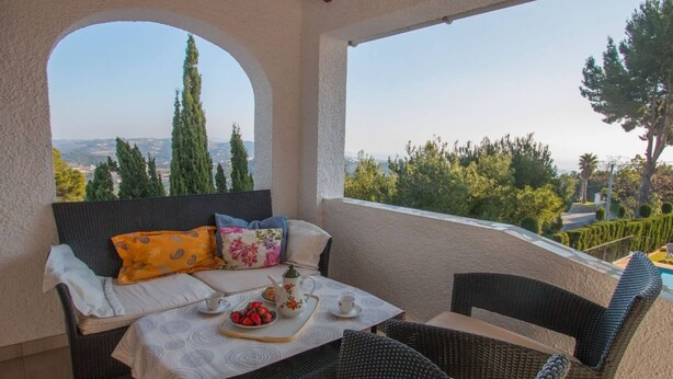 A balcony with wicker furniture, a table with a tray of strawberries, and a view of trees and hills.