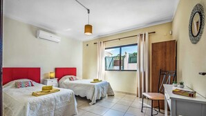A hotel room with two beds, a desk, a chair, a window with curtains, and an air conditioning unit.