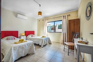 A hotel room with two beds, a desk, a chair, a window with curtains, and an air conditioning unit.