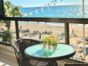 A balcony with a wicker chair and a small table with a glass top. There is a view of a beach with people and umbrellas in the background.