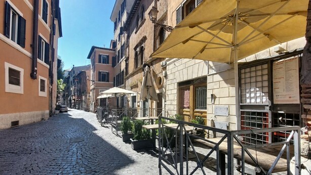 Une rue pavée avec des tables et des chaises installées à l’extérieur, des bâtiments avec des volets et un grand parasol.