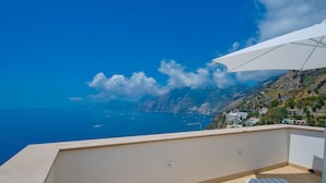 Un balcon avec un parasol blanc, une chaise longue à rayures et une vue sur la mer et les montagnes.