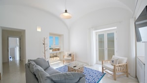 Un salon spacieux avec un canapé gris, une chaise en bois et une table basse en verre. Une grande fenêtre laisse entrer la lumière naturelle et offre une vue sur l’extérieur.