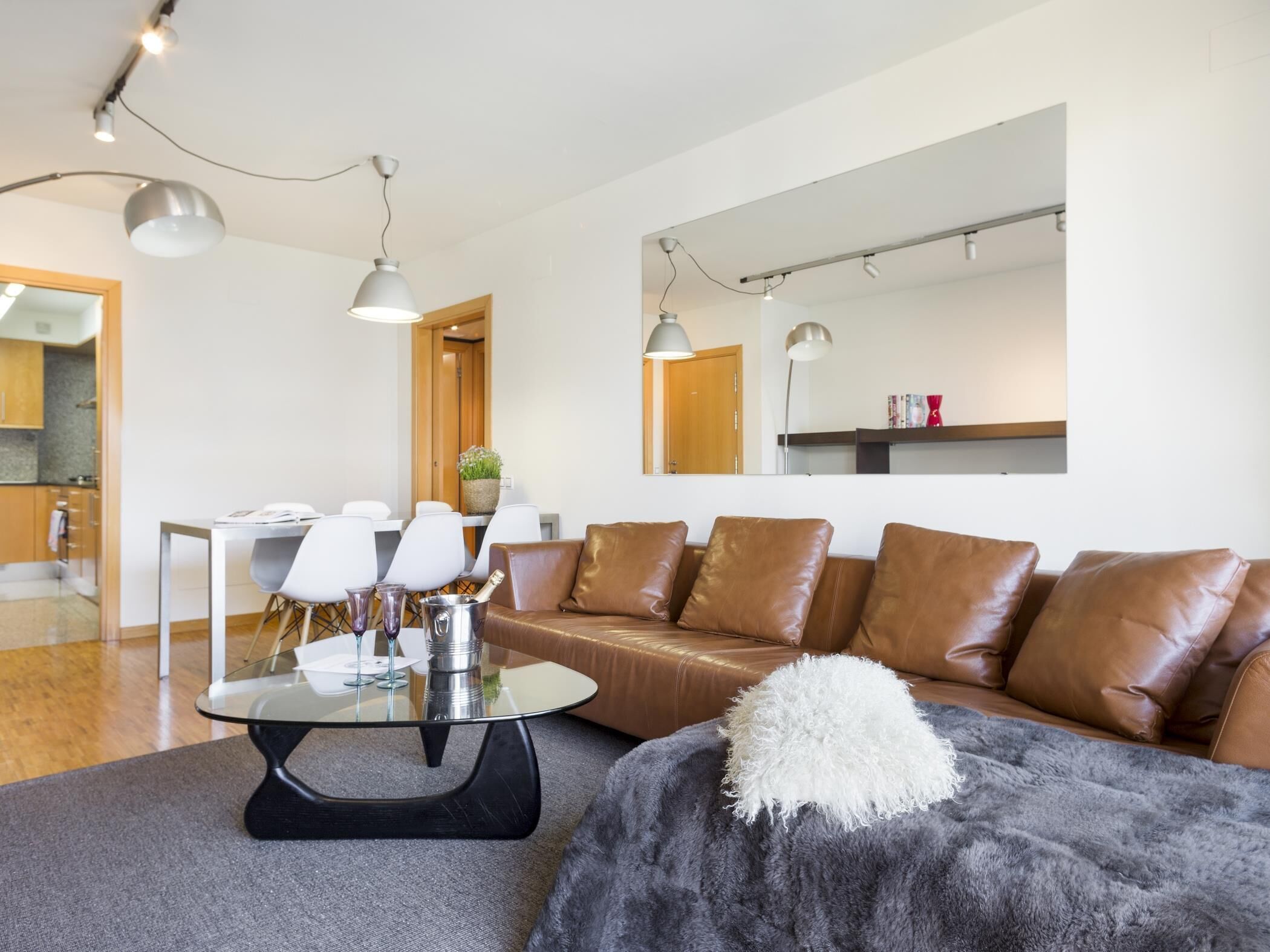 Una sala moderna con un sofá de cuero, una mesa de centro de vidrio y una manta suave. Se observa un comedor con sillas blancas y una pequeña mesa al fondo.