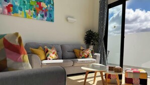 A living room with a grey sofa, colorful cushions, a white coffee table, and a view through a glass door to the outside.