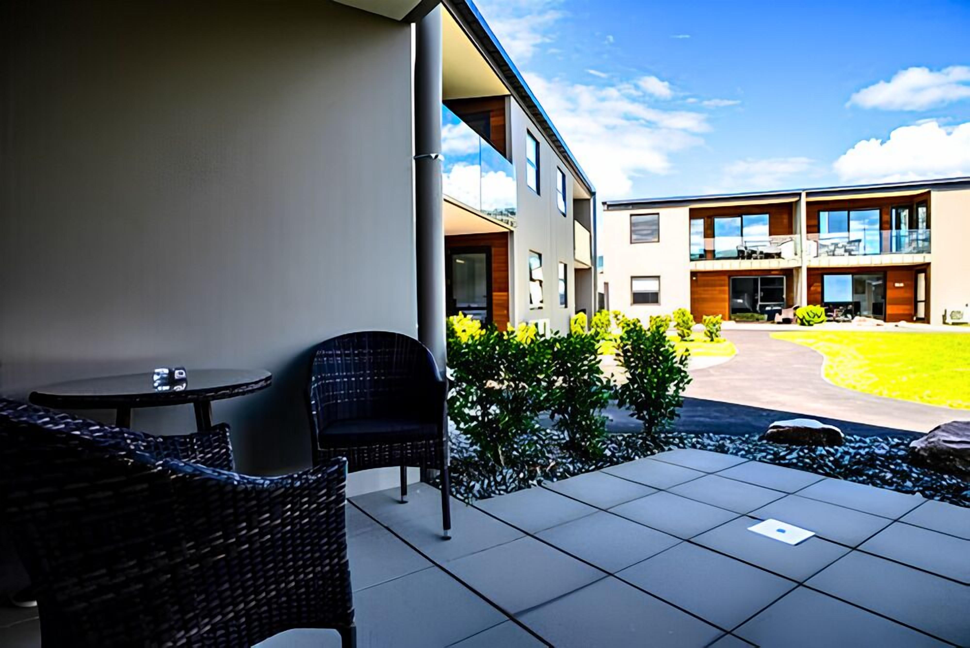An outdoor patio area with a table and chairs, a small garden, and a modern building in the background.