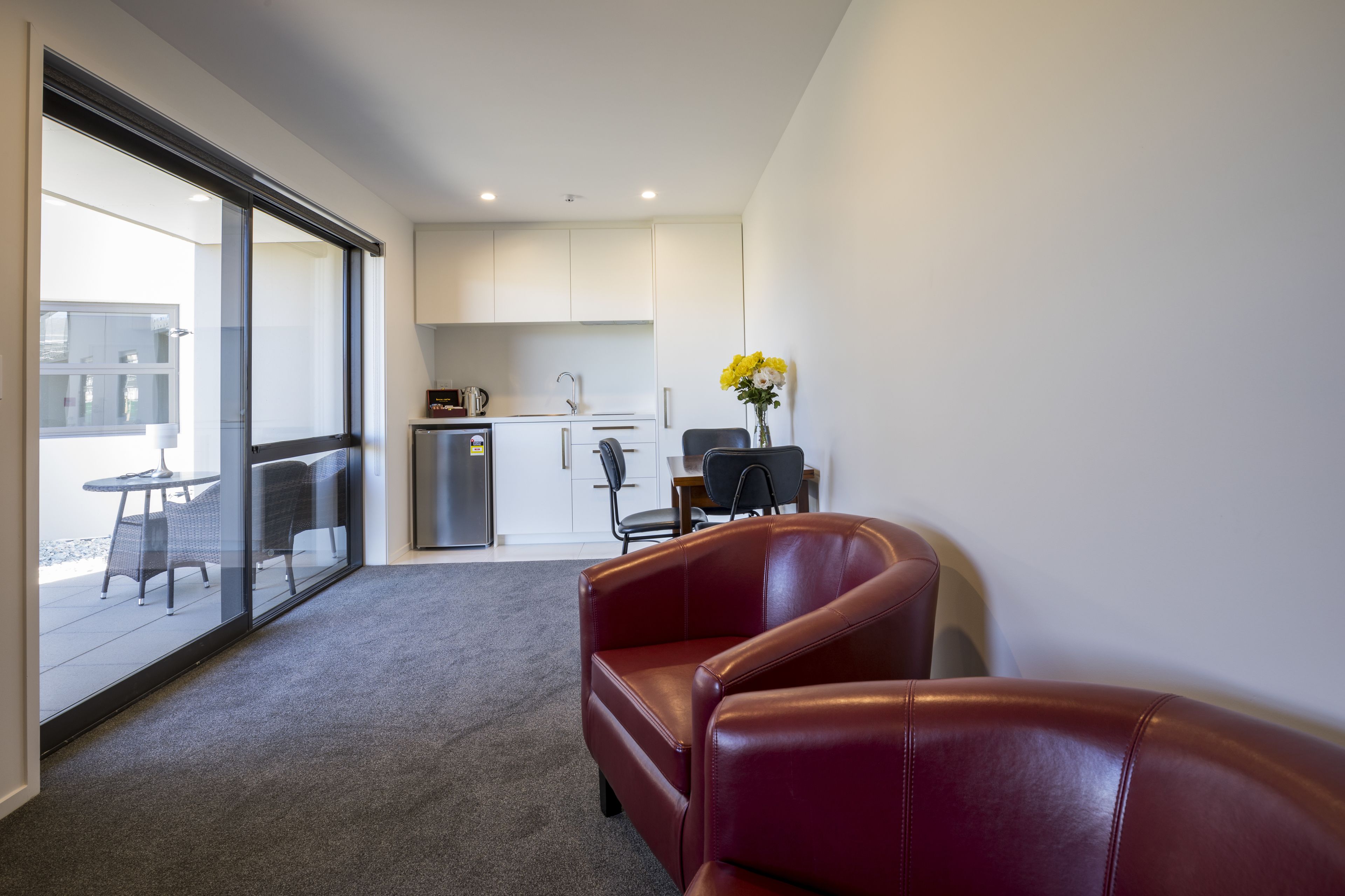Et moderne hotelværelse med en glas skyde dør til en balkon, en rød lædersofa, et lille tekøkken og et spisebord med stole.