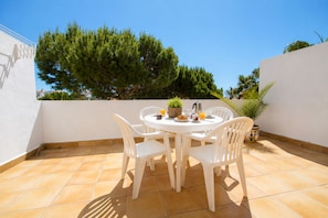 A round table with white chairs, a potted plant, and a kettle on a tiled patio.