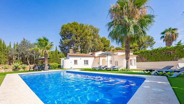 Una piscina con cubierta de baldosas blancas, rodeada de palmeras y una casa con techo a ladrillo.