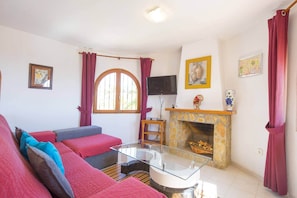 A living room with a sofa set, a glass coffee table, a fireplace, and a TV mounted on the wall. - Villa Cactus - PlusHolidays (Calpe)