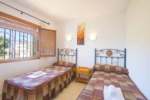 A room with two single beds, a wooden table, and a window with metal bars. - Villa Cactus - PlusHolidays (Calpe)